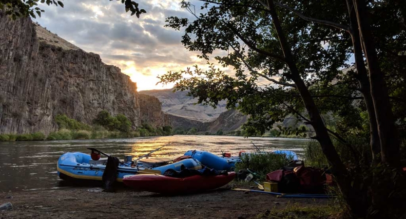 Rafts and kayaks rest on the shore of a calm river, In the background, the river is framed by high canyon walls.